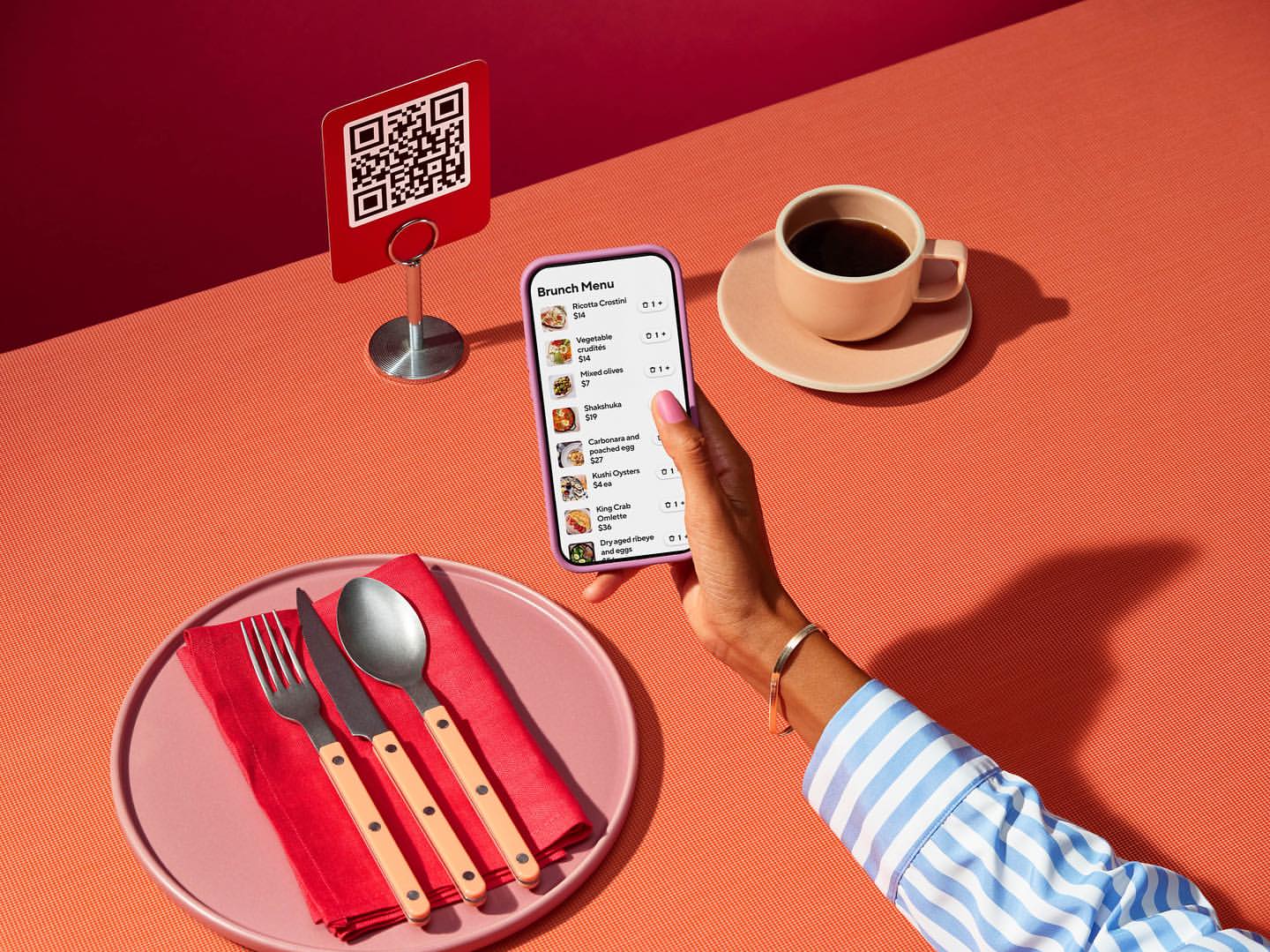 Woman using smartphone to view brunch menu with QR code, coffee, place setting on coral tabletop in brightly lit indoor scene.