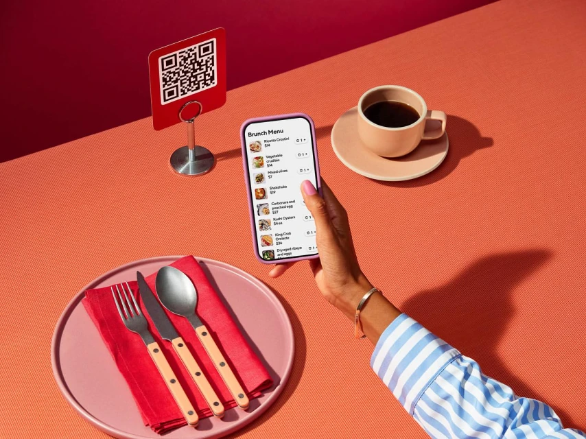 Woman using smartphone to view brunch menu with QR code, coffee, place setting on coral tabletop in brightly lit indoor scene.