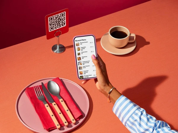 Woman using smartphone to view brunch menu with QR code, coffee, place setting on coral tabletop in brightly lit indoor scene.