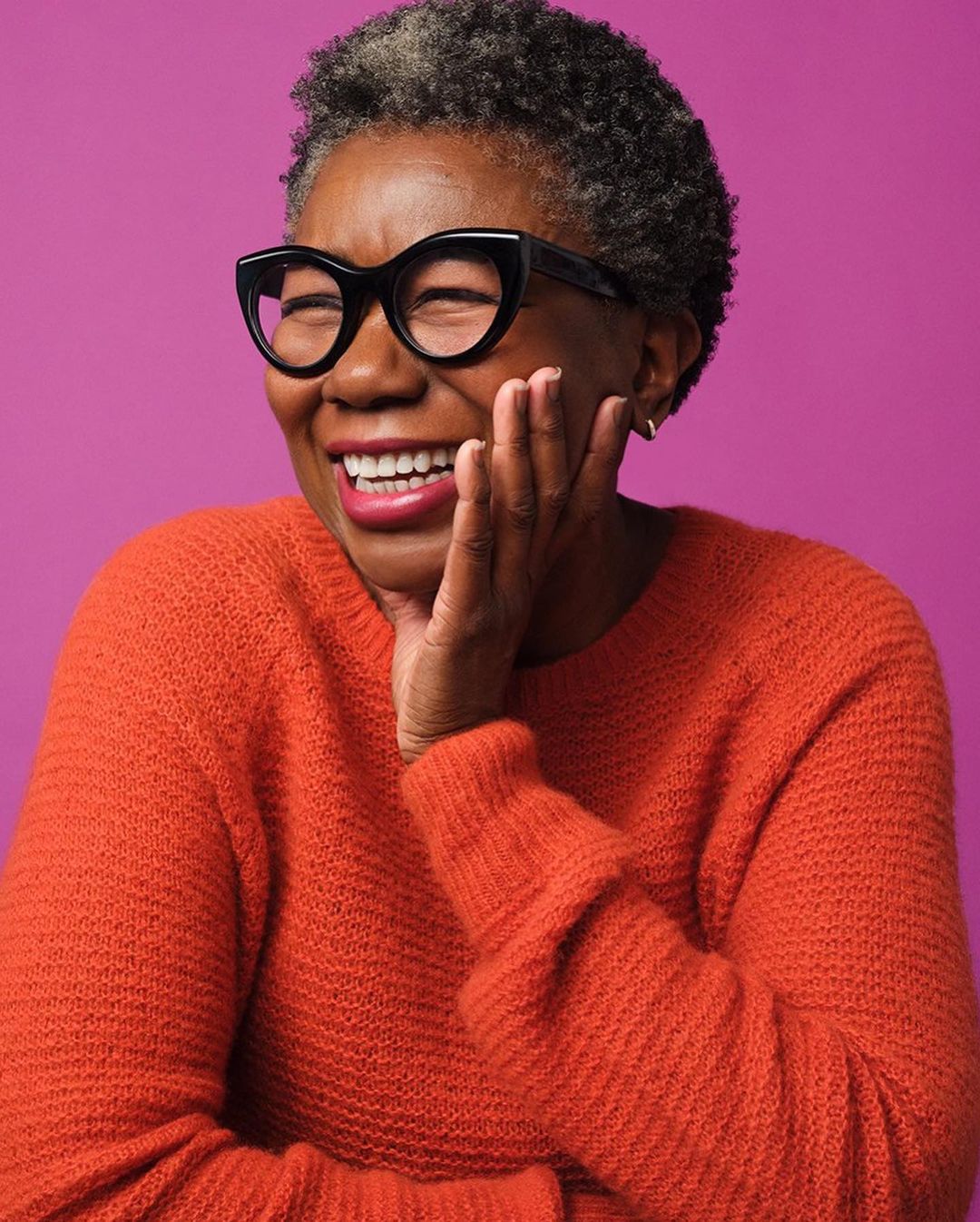 A happy mature woman with short grey hair and black glasses smiles, wearing an orange sweater against a pink background.