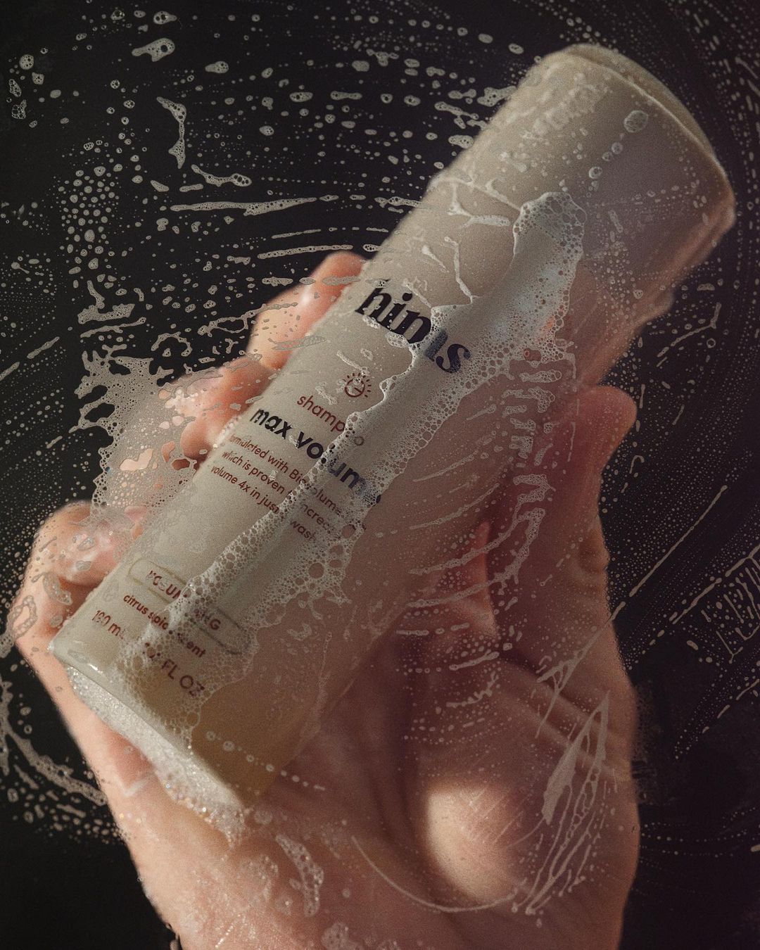 Hand holding a bottle of Hinus shampoo covered in soap bubbles against a dark background in a bathroom setting, close-up.