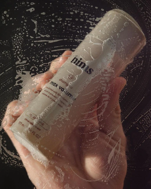 Hand holding a bottle of Hinus shampoo covered in soap bubbles against a dark background in a bathroom setting, close-up.
