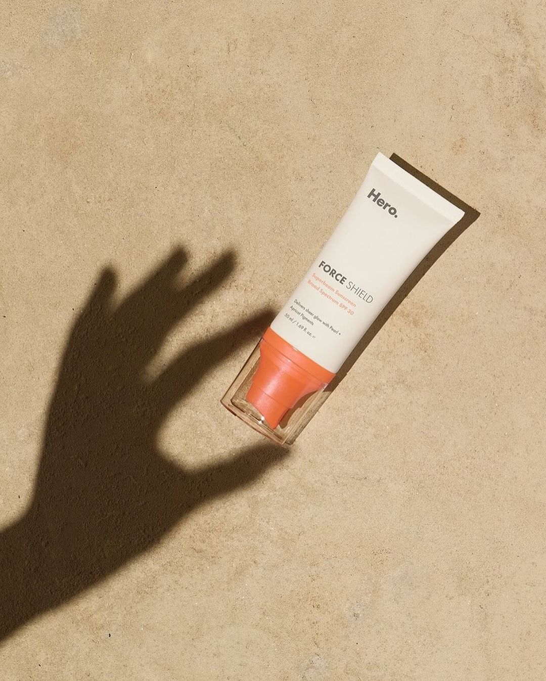 Hero Force Shield sunscreen tube laying on tan surface with hand shadow; product shot with beauty and skincare focus.