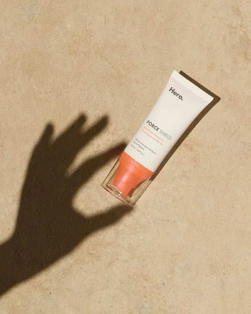 Hero Force Shield sunscreen tube laying on tan surface with hand shadow; product shot with beauty and skincare focus.