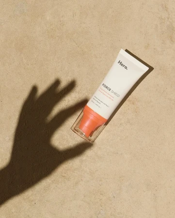 Hero Force Shield sunscreen tube laying on tan surface with hand shadow; product shot with beauty and skincare focus.