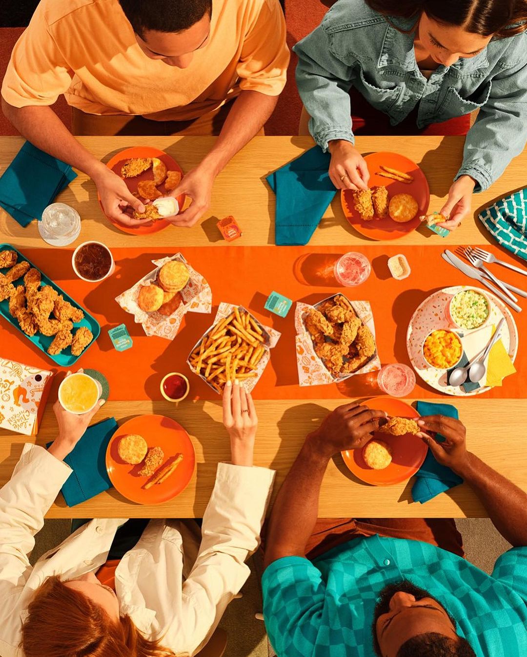 Overhead shot of four people at a wooden table, eating fried chicken, fries, and biscuits, with sauces and drinks, bright orange and blue colors.