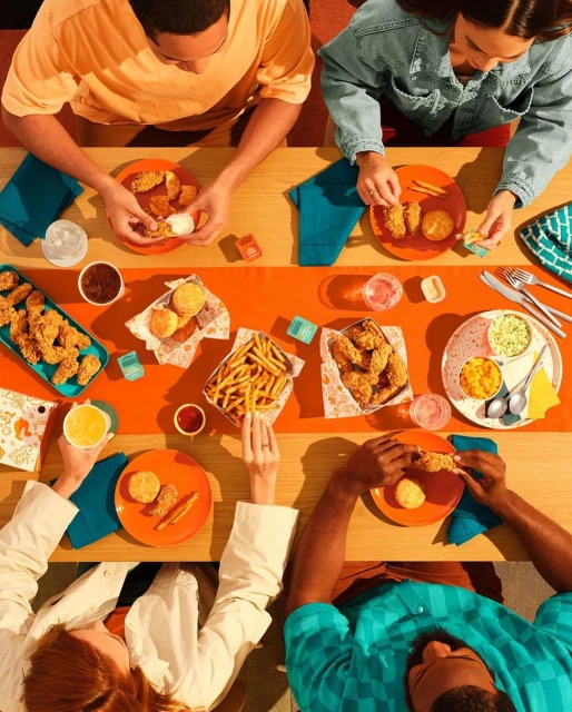 Overhead shot of four people at a wooden table, eating fried chicken, fries, and biscuits, with sauces and drinks, bright orange and blue colors.
