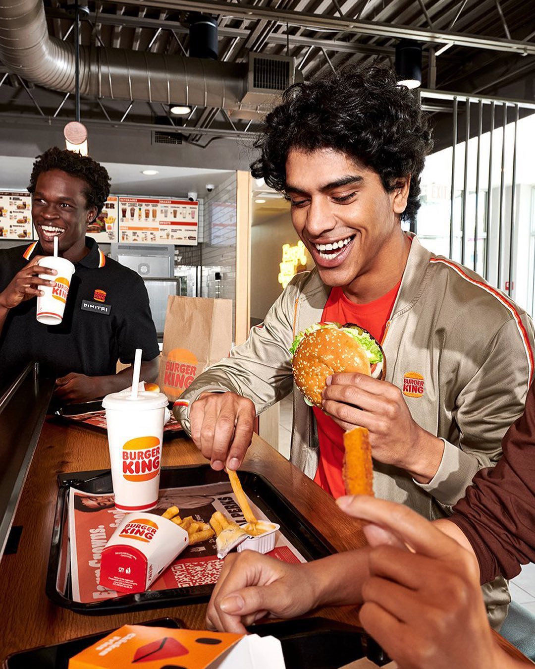 Two smiling men at a Burger King, one holding a burger, the other holding a drink. Fast food items visible on a table.