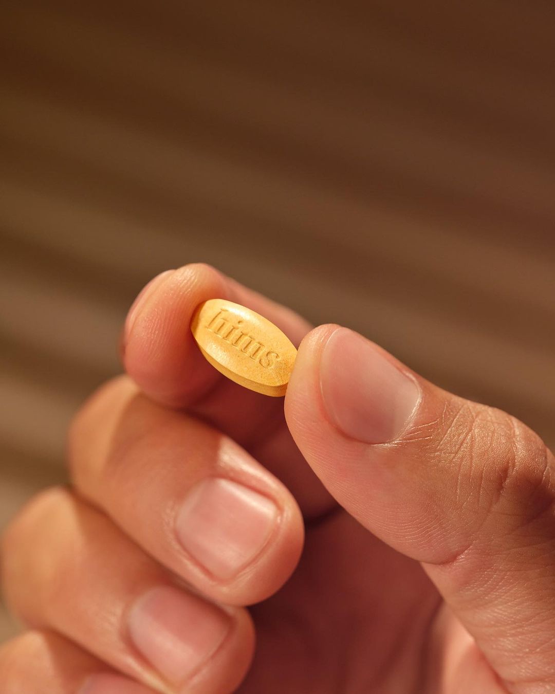 Close-up of a hand holding a single, small, yellow-orange oblong pill with the embossed text 'hims' in a simple, well-lit indoor setting.