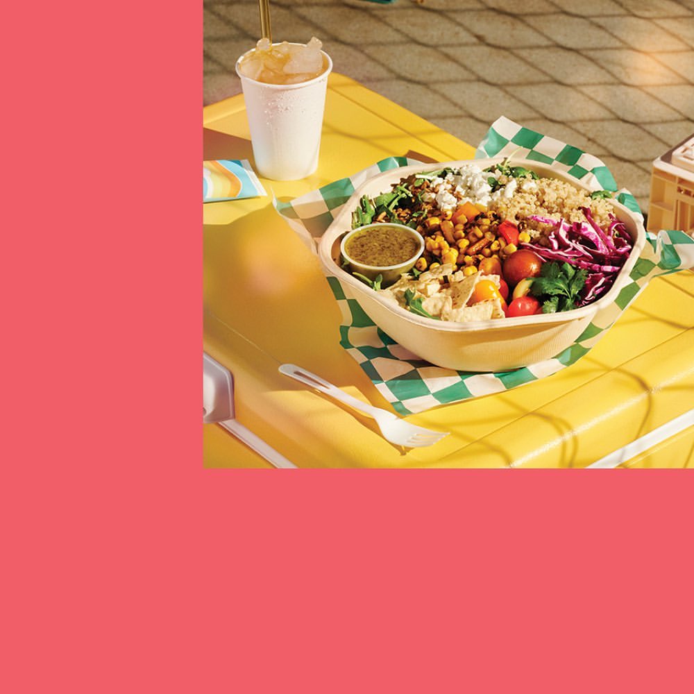 A vibrant salad bowl with tomatoes, grains, and various vegetables on a yellow table, served with a drink in a white paper cup.