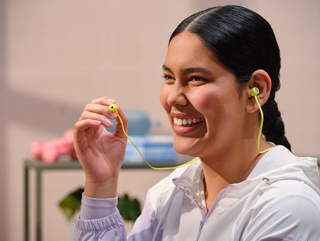 Woman smiles wearing lime-green Beats wireless earbuds. Indoor shot with soft background. Hand near earbud. Activewear is visible.