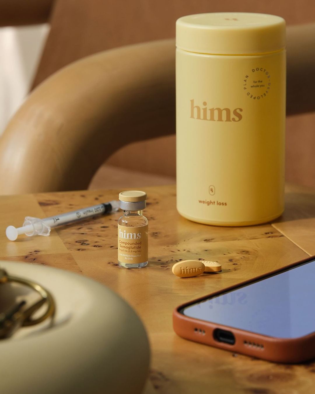 Hims product shot: yellow bottle labeled 'weight loss,' syringe, small vial, pills, and smartphone on a textured tan surface.
