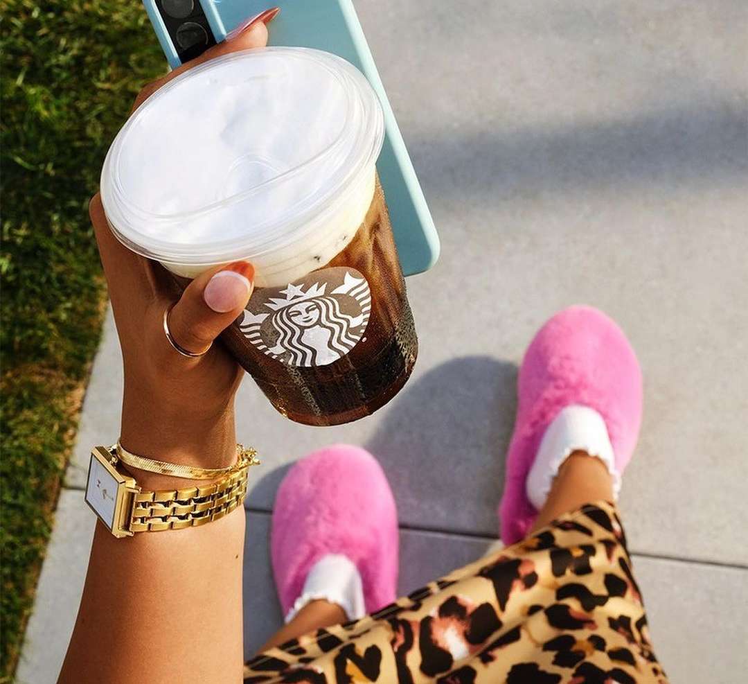 Woman holds iced coffee in a plastic cup in natural light, wearing pink slippers and a gold watch, standing outdoors on pavement.