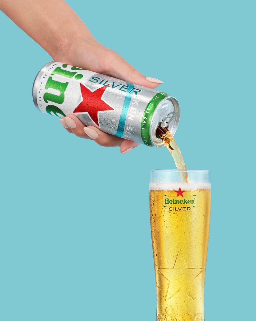 Close-up of a hand pouring Heineken Silver beer from an aluminum can into a tall glass on a bright turquoise background.