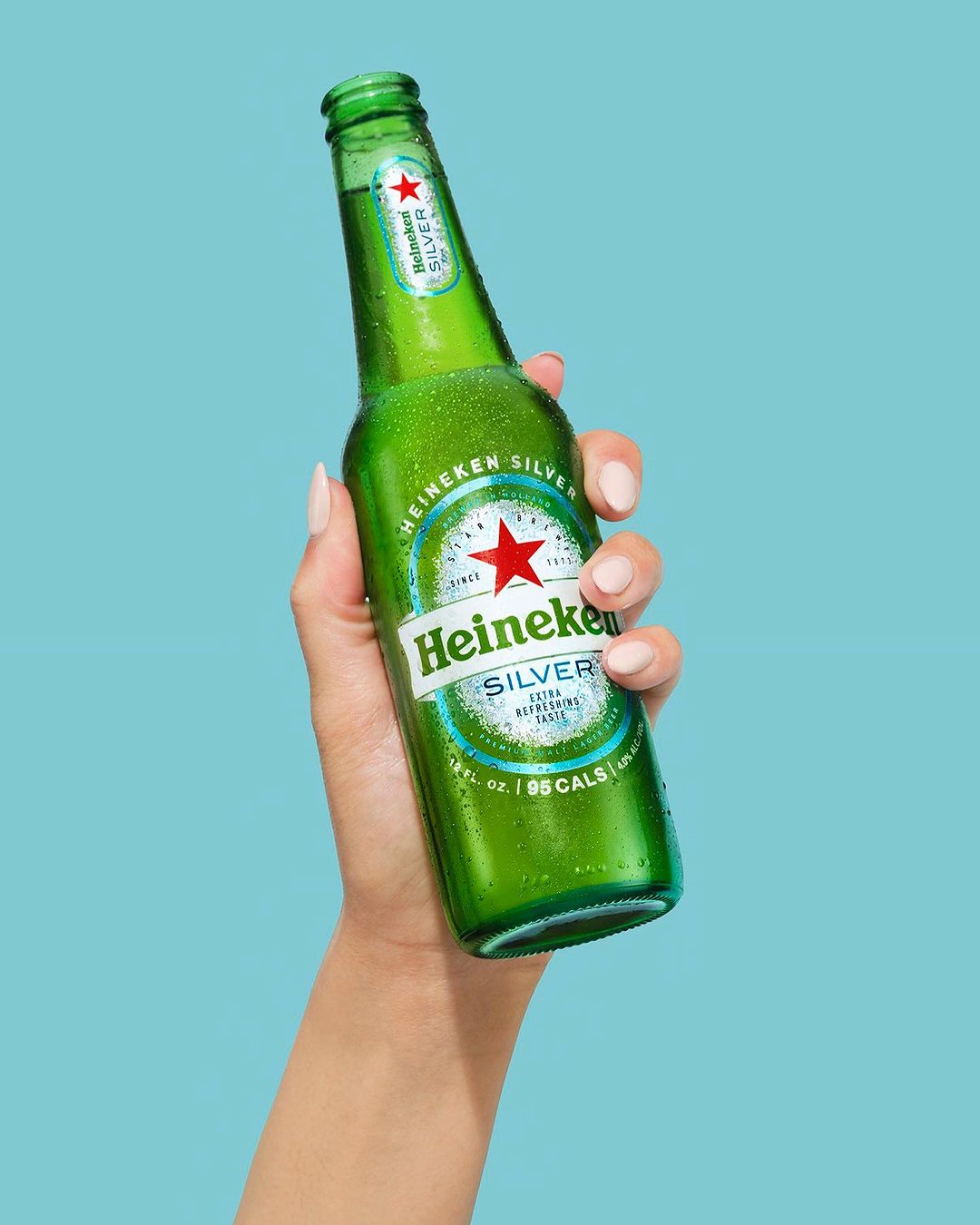Hand holding a green Heineken Silver beer bottle against a light blue background in a studio shot promoting beverage freshness.