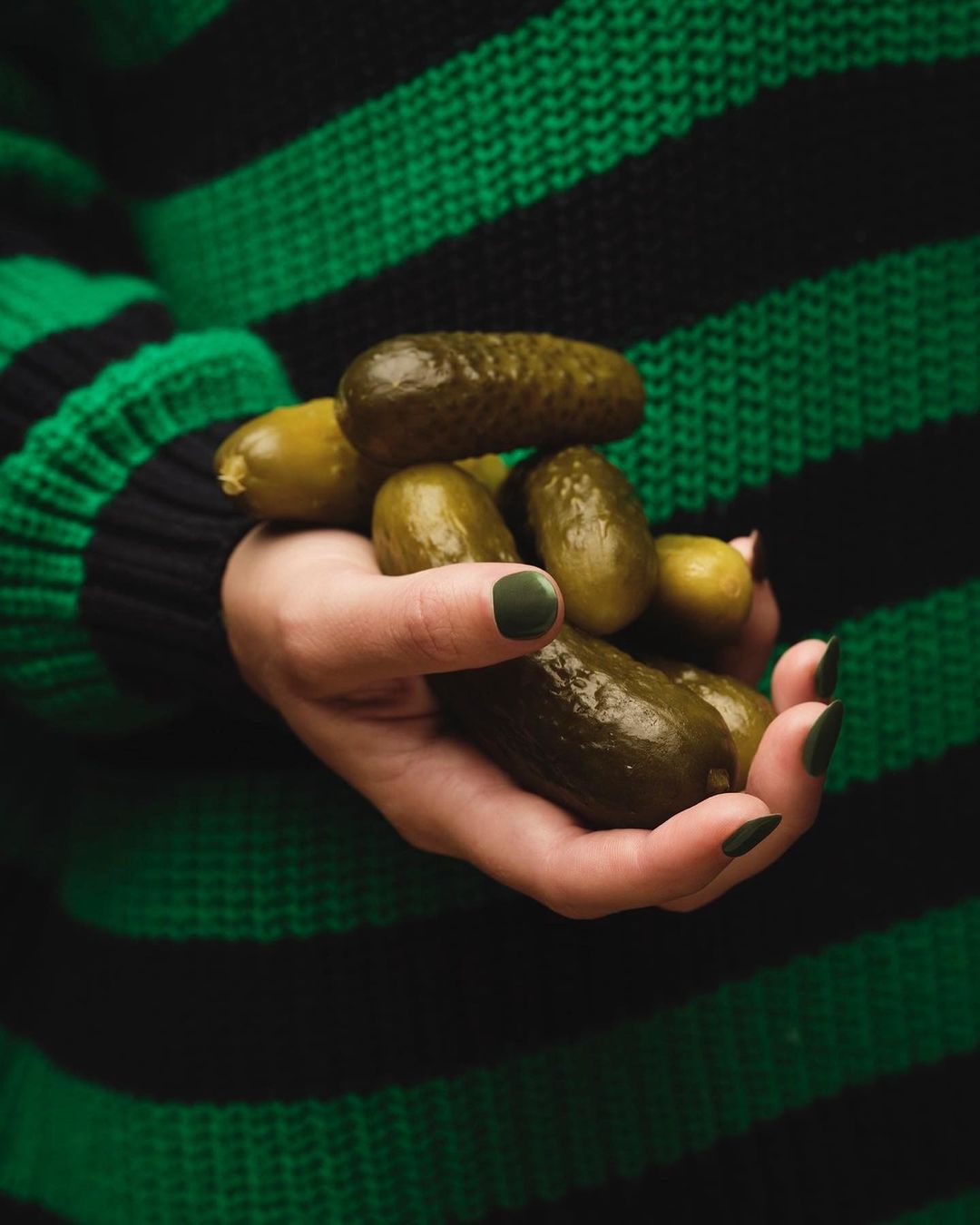Hand holding a pile of green pickles against a dark green and black striped sweater in a close-up studio food shot.
