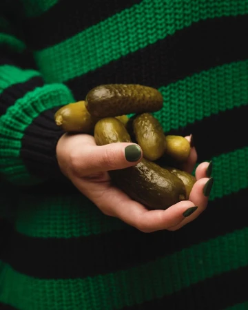 Hand holding a pile of green pickles against a dark green and black striped sweater in a close-up studio food shot.