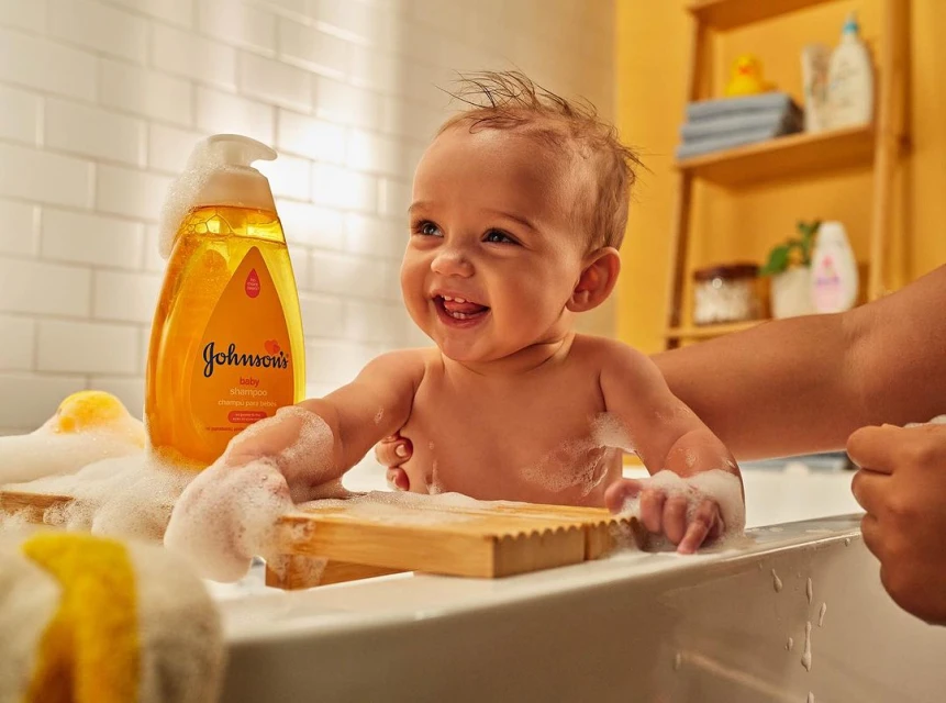 Happy baby smiles in a bath with Johnson's baby shampoo bottle and soap suds, showcasing a cheerful bath time scene.