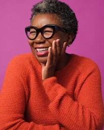 Close-up portrait of an elderly Black woman smiling in a bright orange textured sweater and black cat-eye glasses against a solid pink background.