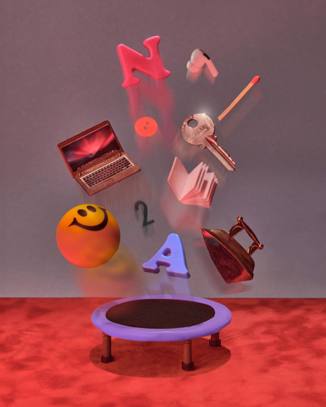 Still life composition featuring floating letters, a laptop, book, smiley face ball, and other objects over a trampoline.