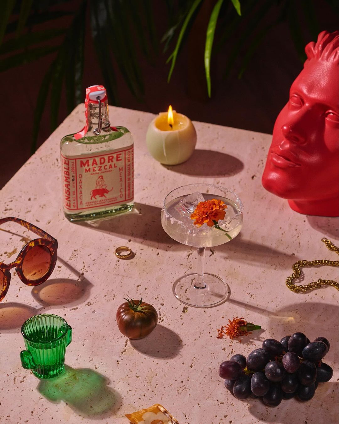 Still life shot with a bottle of Madre Mezcal, candle, cocktail glass with flower, grapes, and a bust in a warm-toned studio.