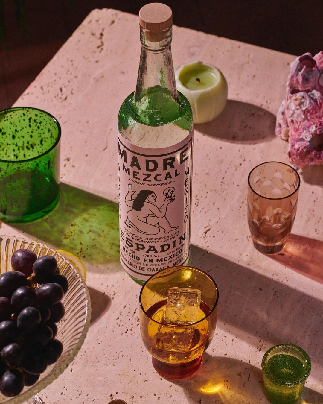 A bottle of Madre Mezcal mezcal surrounded by colored glassware, grapes, a lemon wedge, and a lit candle against a textured surface.