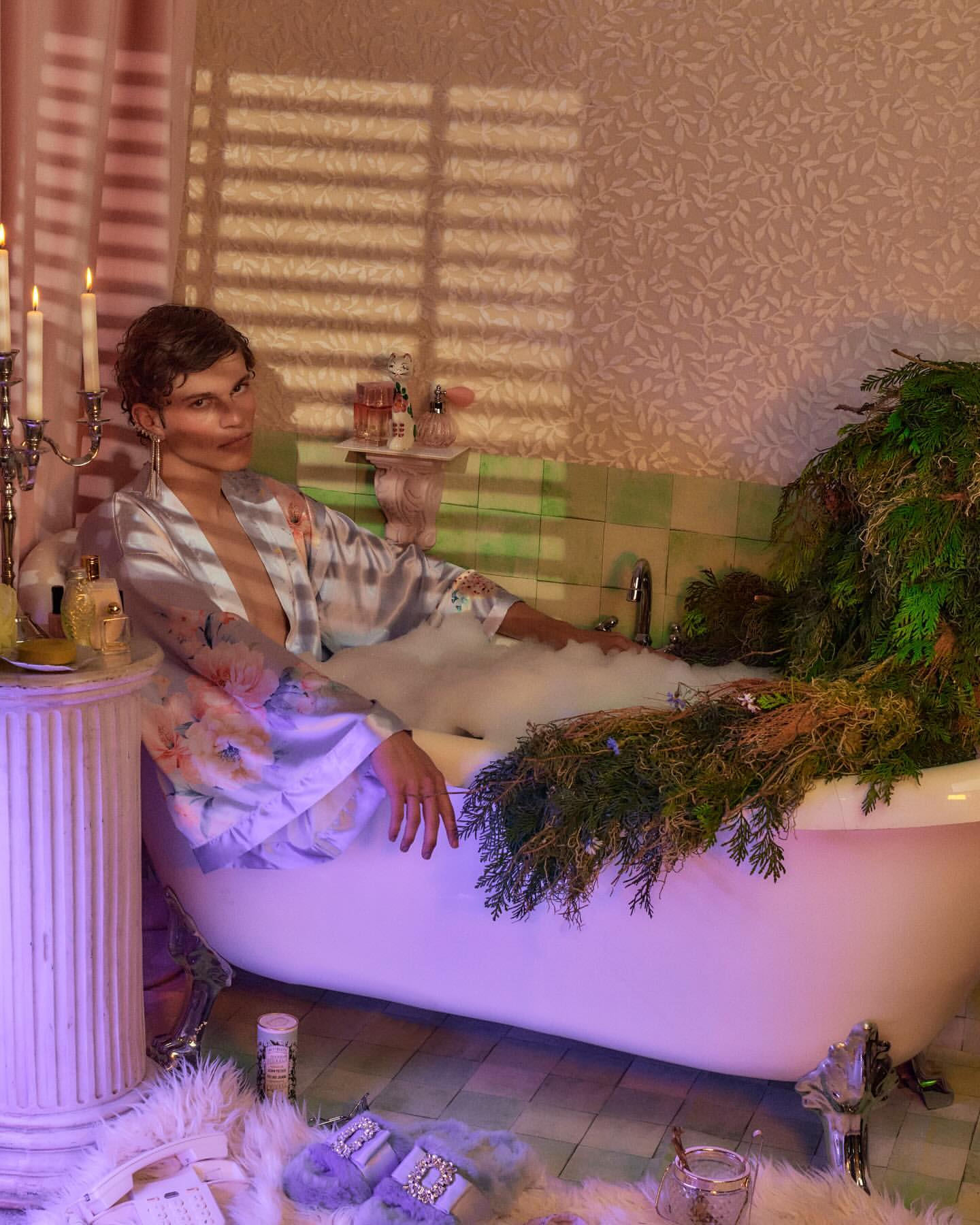 Woman in a floral robe reclines in a clawfoot bathtub filled with bubbles, accessorized with slippers and a column-style side table.