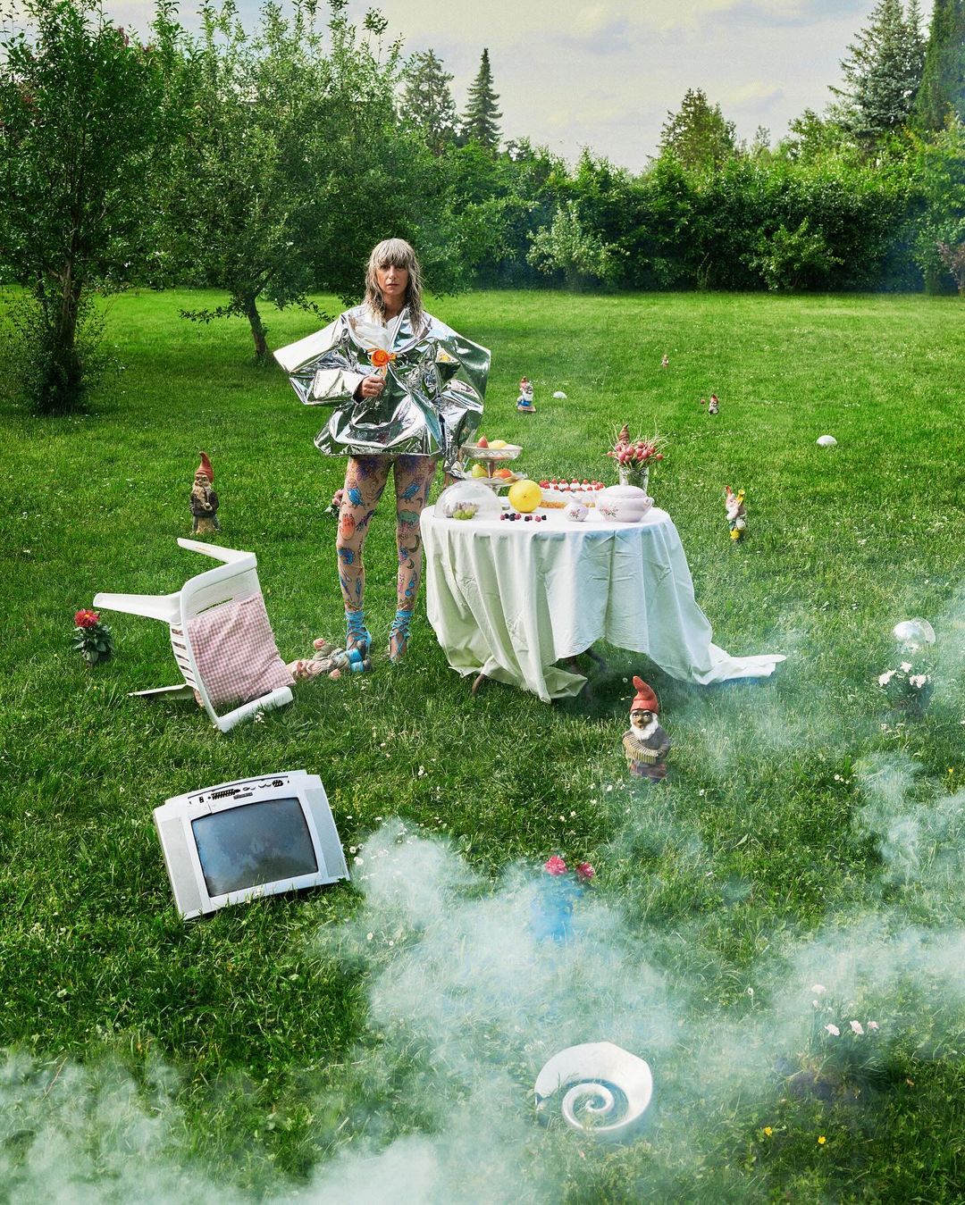Woman in futuristic silver outfit stands in grassy yard with vintage TV, garden gnomes, and white table with snacks under cloudy skies.