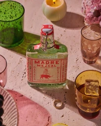 Madre Mezcal bottle surrounded by colorful glassware, a lit candle, and ice on a textured stone surface