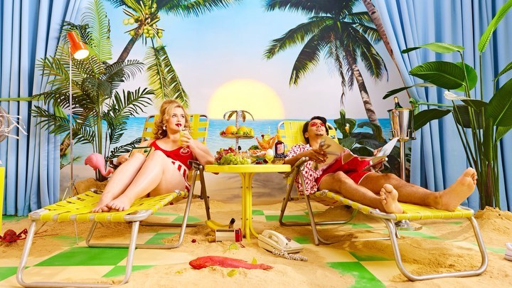 Retro beach scene with a man and woman lounging by a table with fruit and drinks against a painted backdrop of a beach landscape.