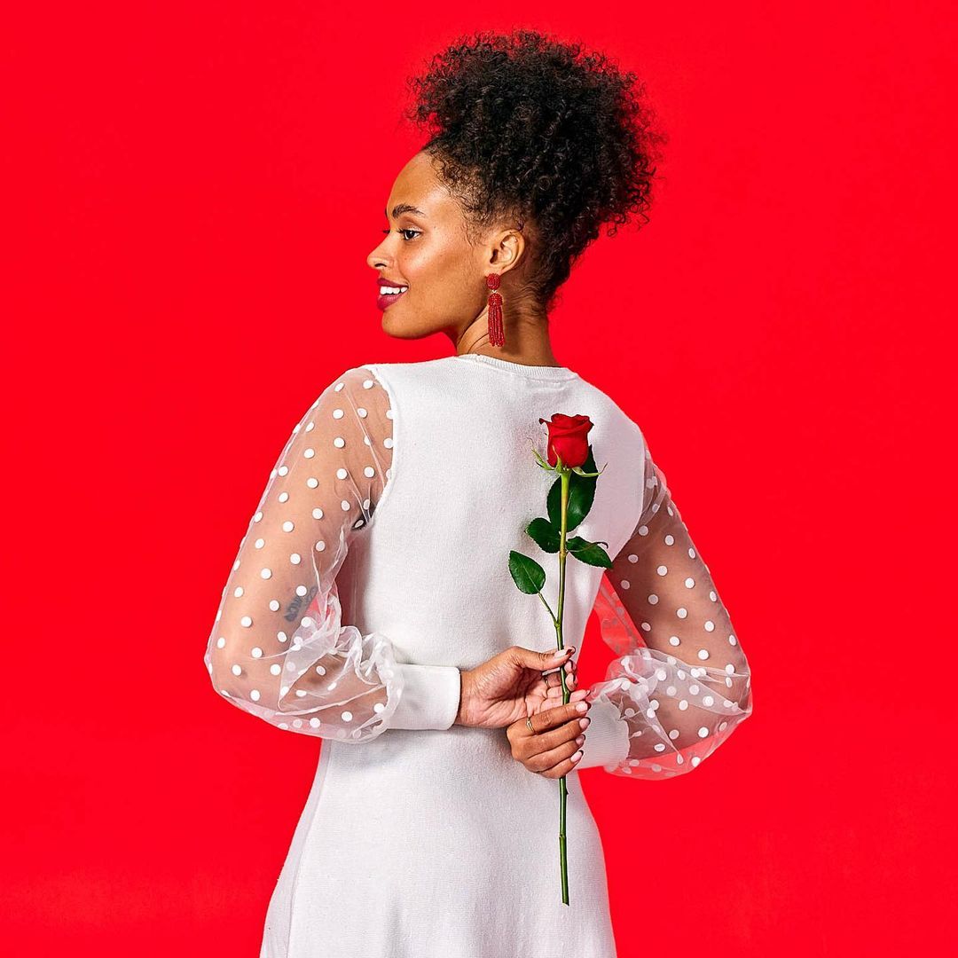 Woman with dark hair smiles in profile, wearing a white dress, holding a single red rose behind her back, against a red background.