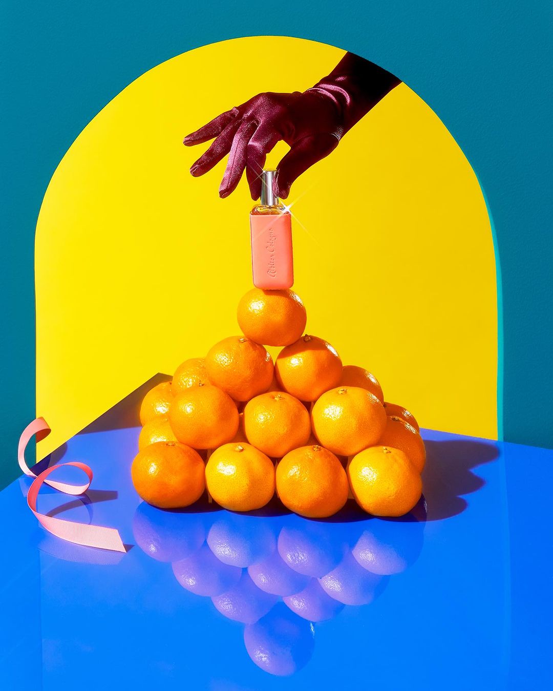 Gloved hand places perfume bottle on a pyramid of oranges against a blue and yellow background with pink ribbon accent.