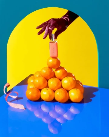 Gloved hand places perfume bottle on a pyramid of oranges against a blue and yellow background with pink ribbon accent.