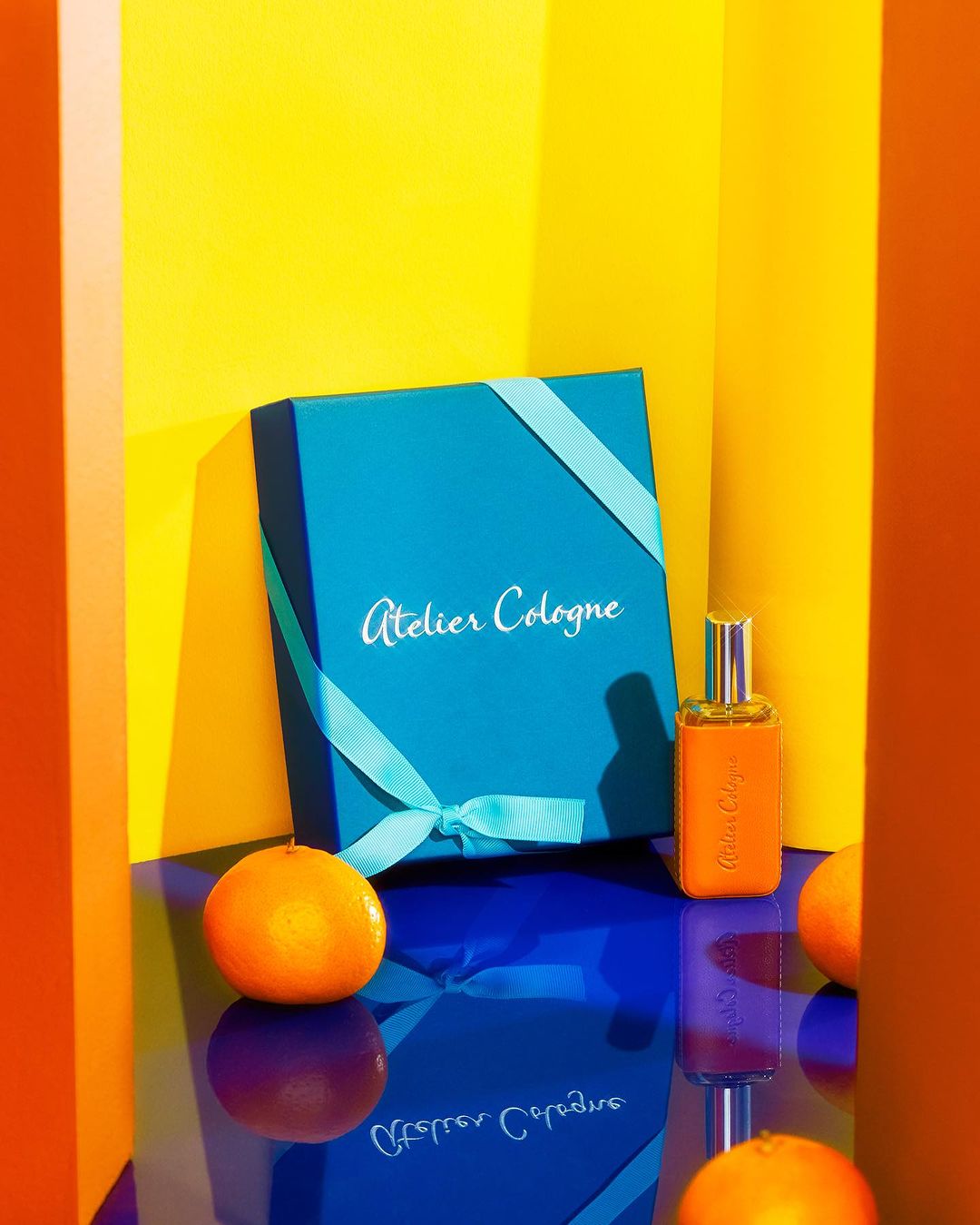 Studio product shot of Atelier Cologne fragrance bottle and turquoise gift box with oranges against bright yellow and blue backdrop.