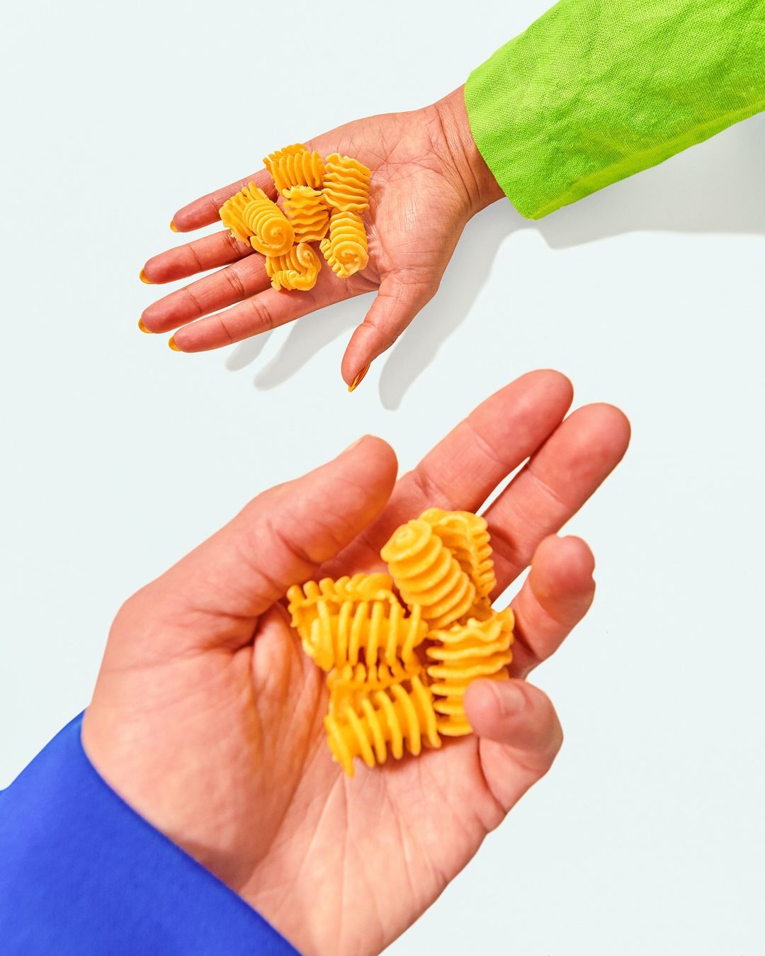 Two hands, one in bright green and one in blue, each holding a pile of rotini pasta on a solid white background with studio lighting.