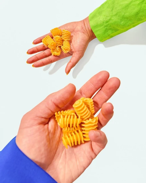 Two hands, one in bright green and one in blue, each holding a pile of rotini pasta on a solid white background with studio lighting.