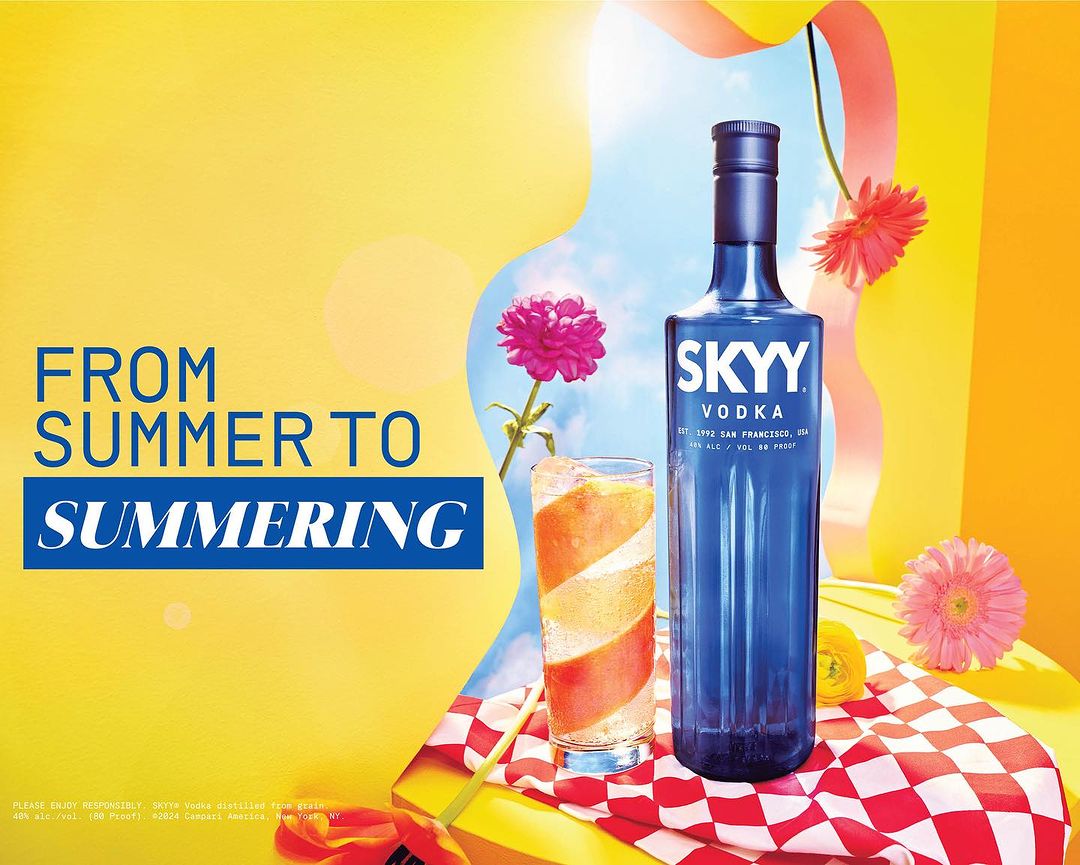 A bottle of Skyy Vodka stands tall next to a cocktail glass, set on a red-and-white checkered cloth, against a vibrant yellow backdrop.