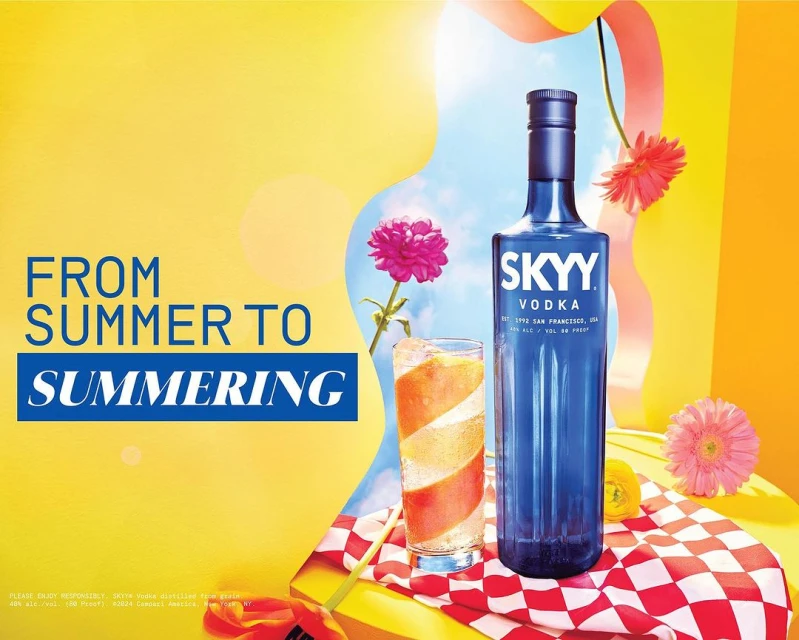 A bottle of Skyy Vodka stands tall next to a cocktail glass, set on a red-and-white checkered cloth, against a vibrant yellow backdrop.