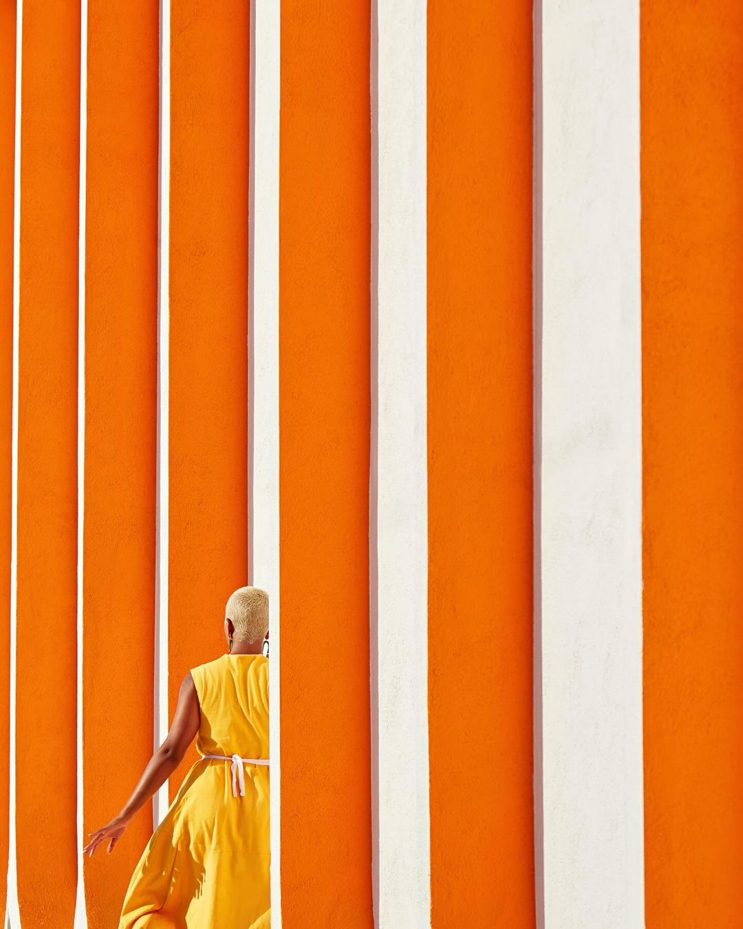 Person in yellow dress walks alongside an orange wall with vertical white stripes, bathed in bright sunlight. Minimalist composition.