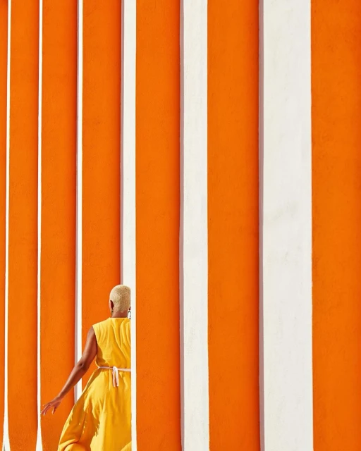 Person in yellow dress walks alongside an orange wall with vertical white stripes, bathed in bright sunlight. Minimalist composition.