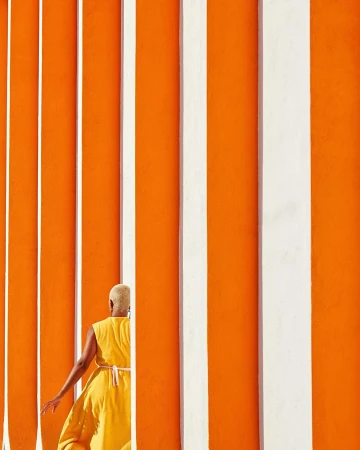 Person in yellow dress walks alongside an orange wall with vertical white stripes, bathed in bright sunlight. Minimalist composition.