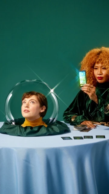 Two women at a table with cards, one in a glass bowl, one holding a cell phone with a nature scene. Teal background, portrait orientation.
