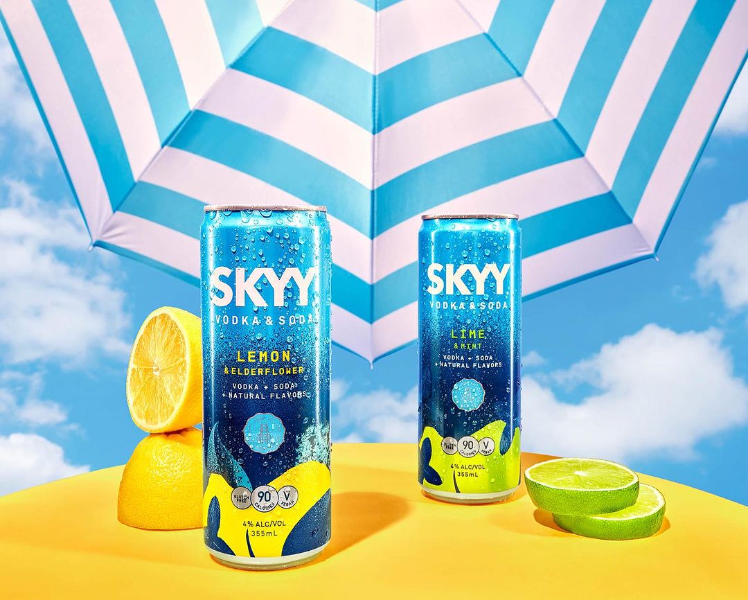 Two cans of Skyy Vodka & Soda stand on a yellow surface with lemons and limes, set against a striped umbrella and blue sky.