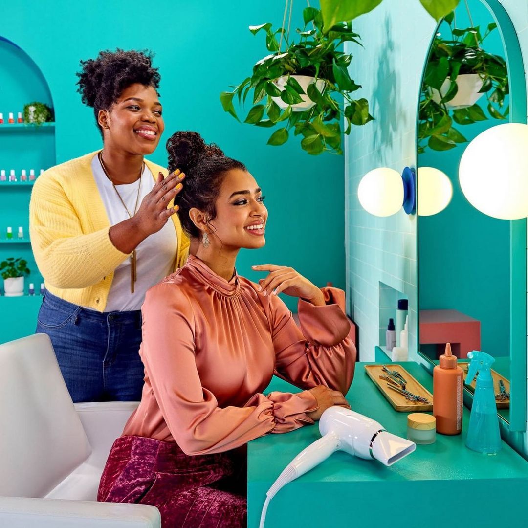 Two women in a hair salon, one standing with hands on the other's hair who is seated, with teal walls, mirror, and hair styling tools.