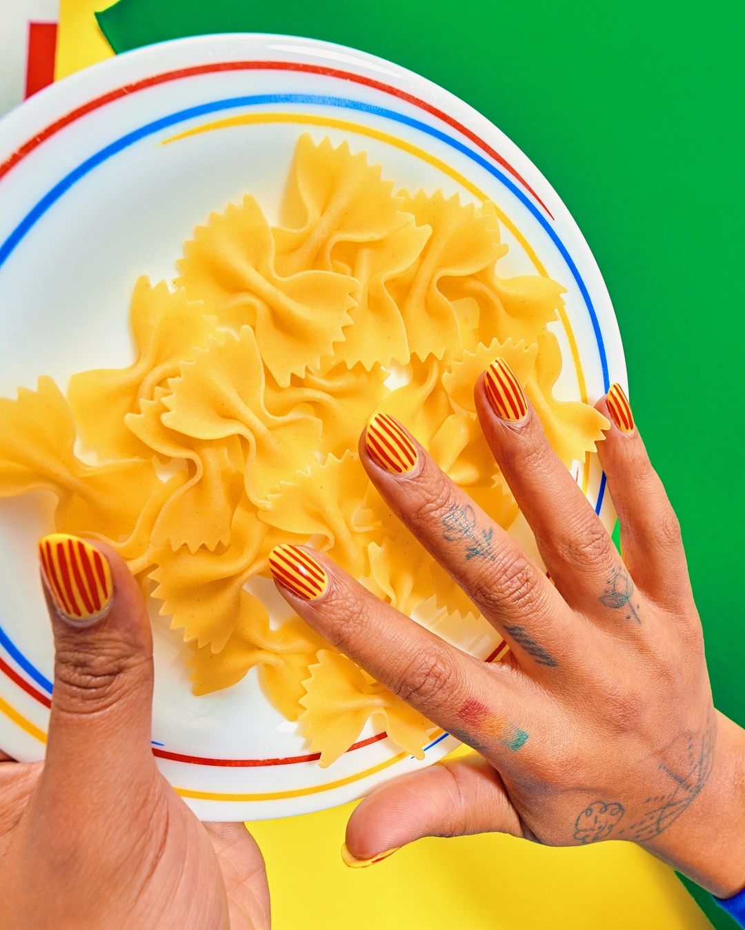 Overhead shot of farfalle pasta on a plate, held by a woman's hands, fingernails are decorated with a striped pattern, vibrant colors.