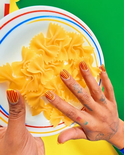 Overhead shot of farfalle pasta on a plate, held by a woman's hands, fingernails are decorated with a striped pattern, vibrant colors.