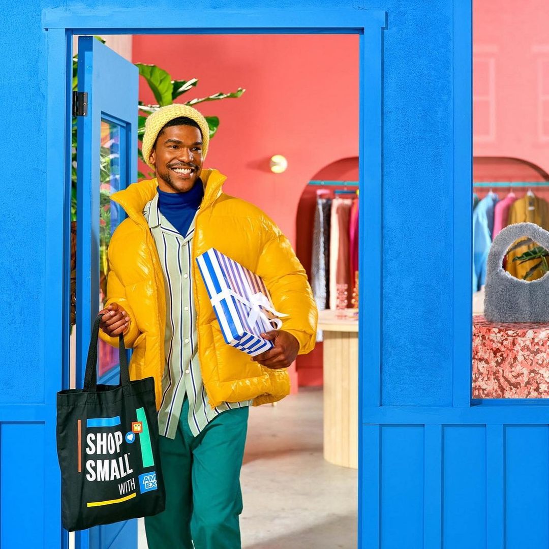 A smiling Black male in a yellow puffer jacket entering a shop, carrying a wrapped gift and tote bag with the words 'Shop Small'.