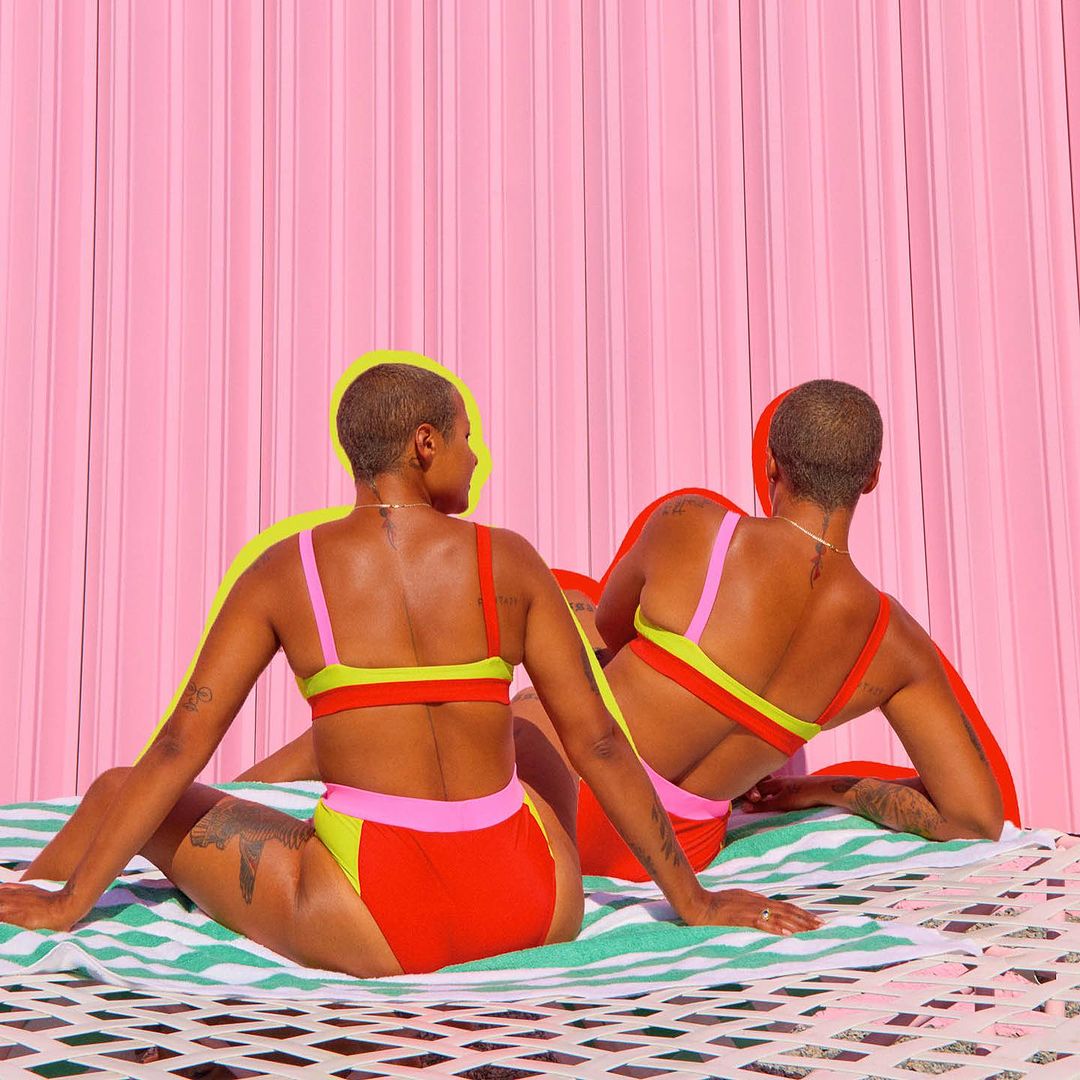 Two women in vibrant swimsuits sit on a striped towel against a pink backdrop, showcasing summer fashion and body positivity in a studio setting.