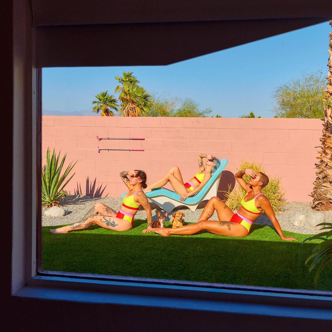Three women in swimsuits relax on lounge chair and grass in a modern courtyard with pink wall under clear blue sky; seen through a window.