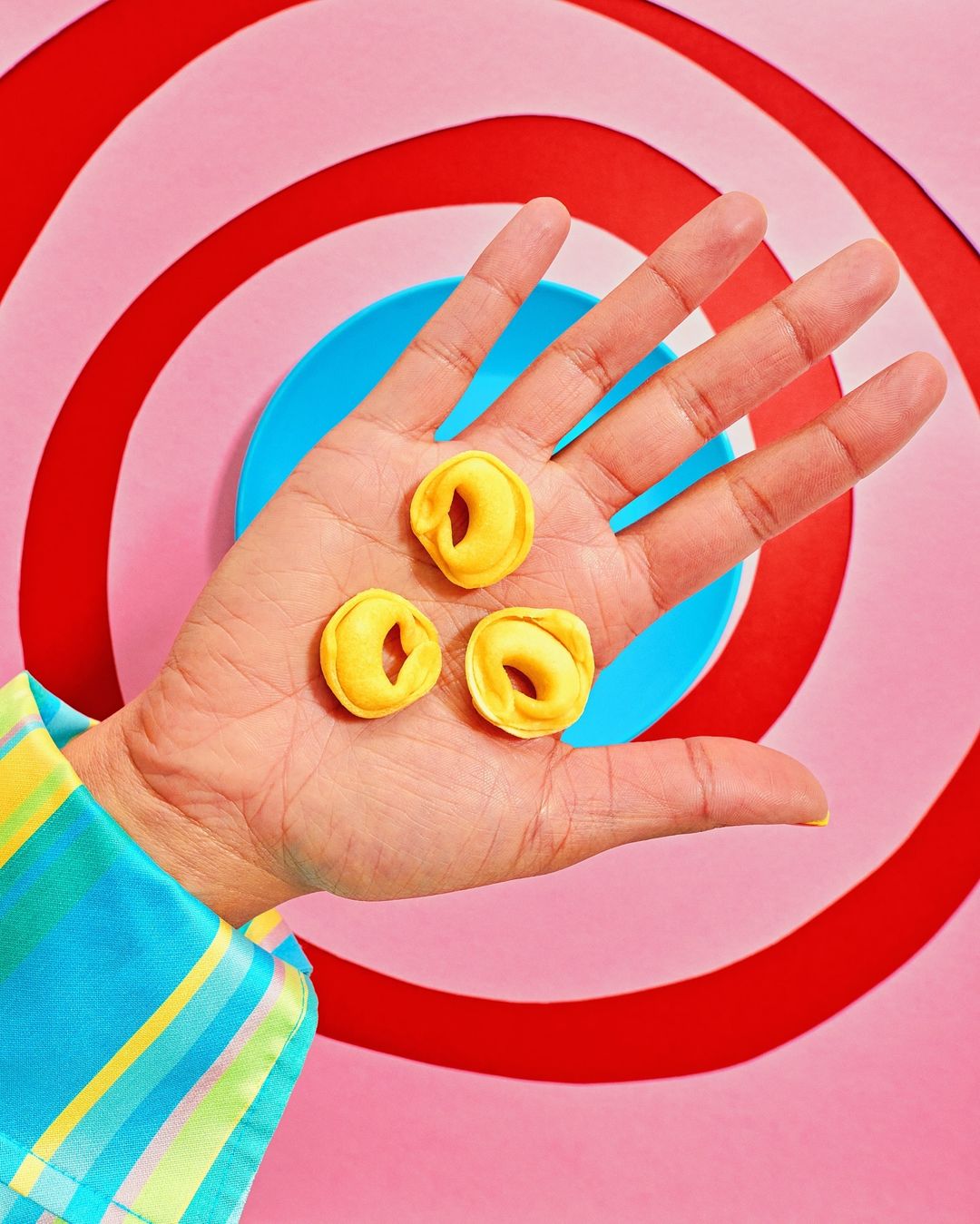 Three ring-shaped pasta pieces are held in an adult's hand against a colorful, graphic background. The sleeve is patterned with squares.